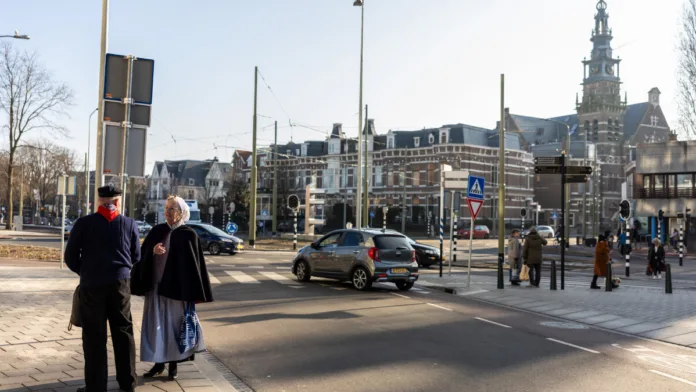 Twee ouderen in traditionele klederdracht praten op een kruising in Scheveningen-Dorp met historische gebouwen op de achtergrond.